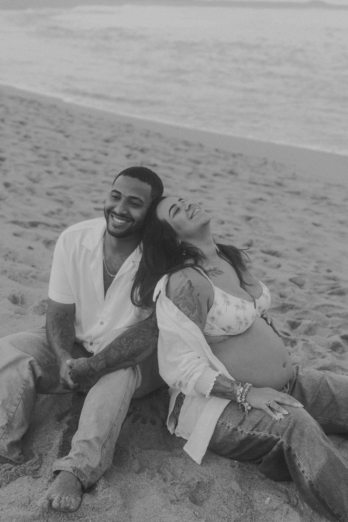 Casal sentados e sorrindo na praia em fotos de gestante.
