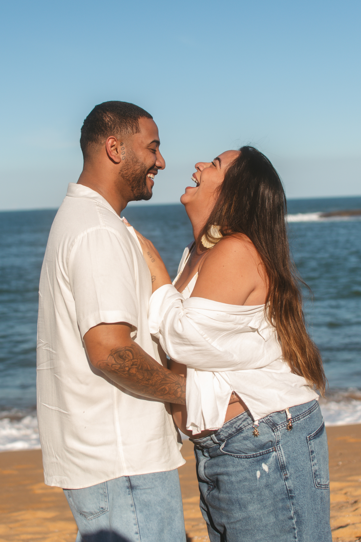 Casal trocando carinho e risadas durante ensaio fotográfico de gestante na praia.