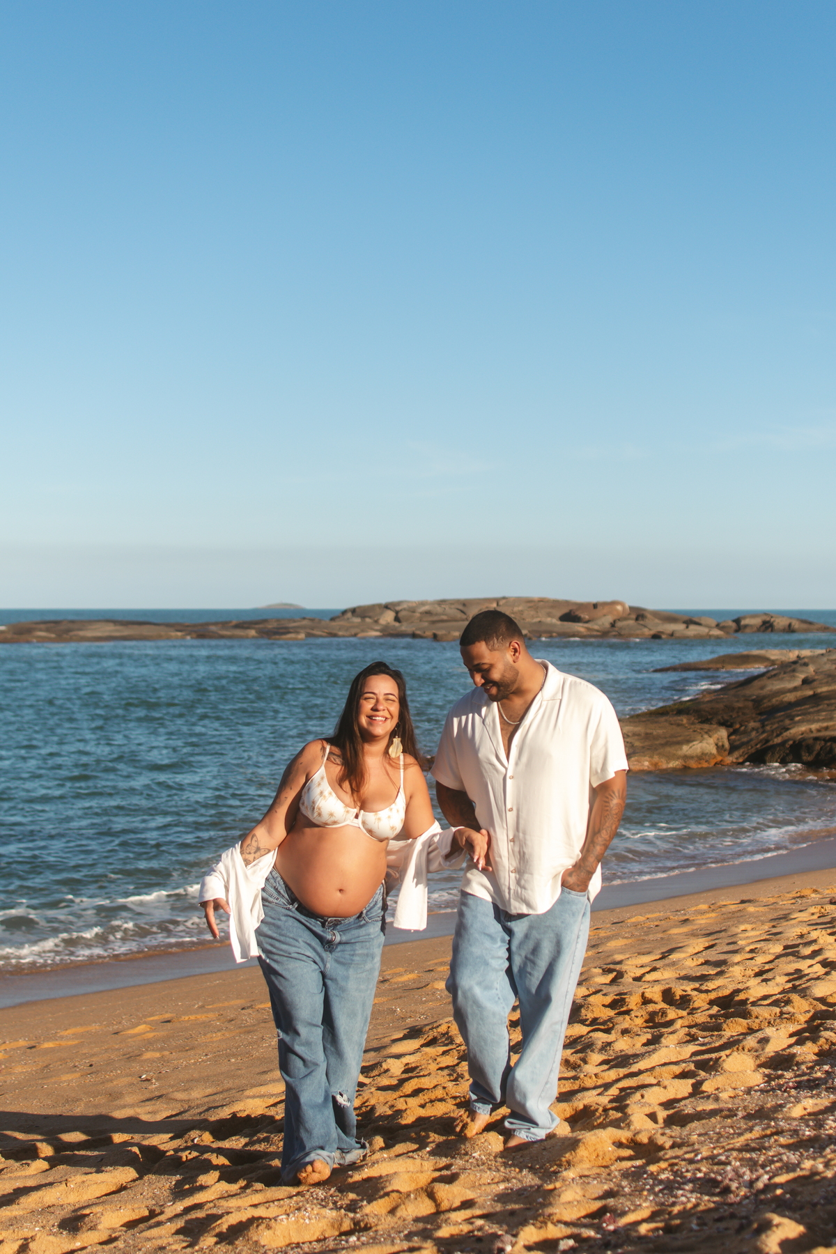 Casal caminhando pela praia durante ensaio de gestante ao entardecer.