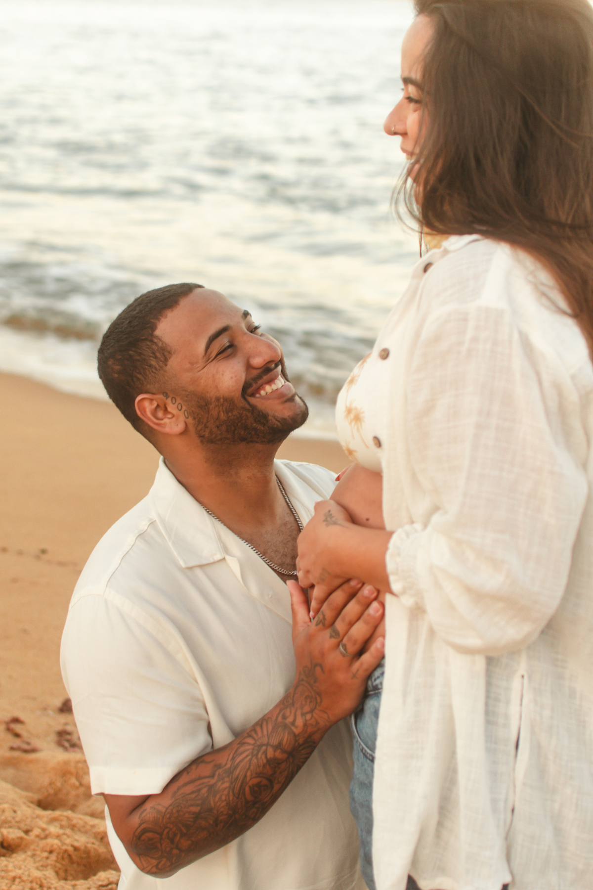 Detalhe de marido sorrindo para esposa grávida durante ensaio na praia.