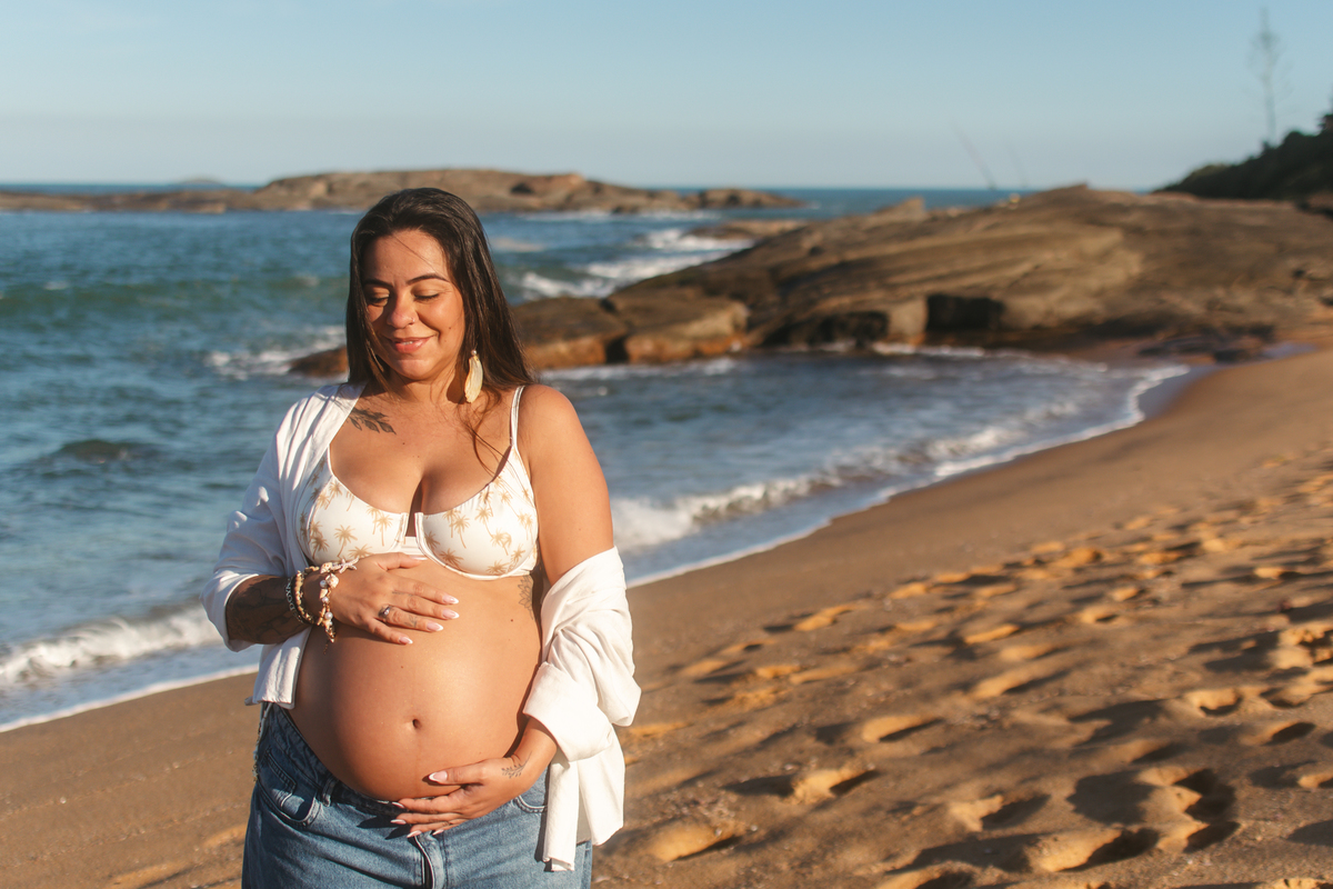 Gestante na praia em Guarapari, mar e pedras ao fundo.