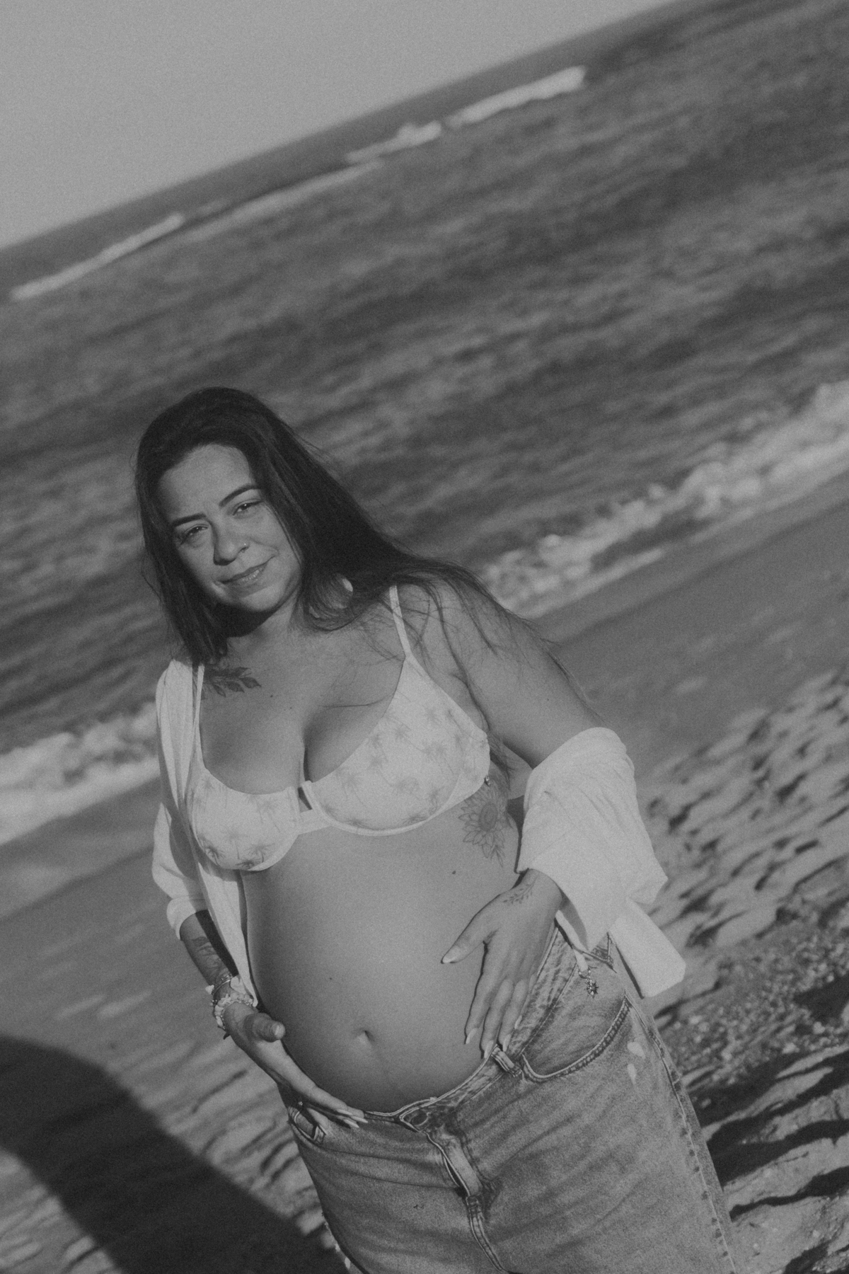 Gestante com mar ao fundo em ensaio na praia em preto e branco.