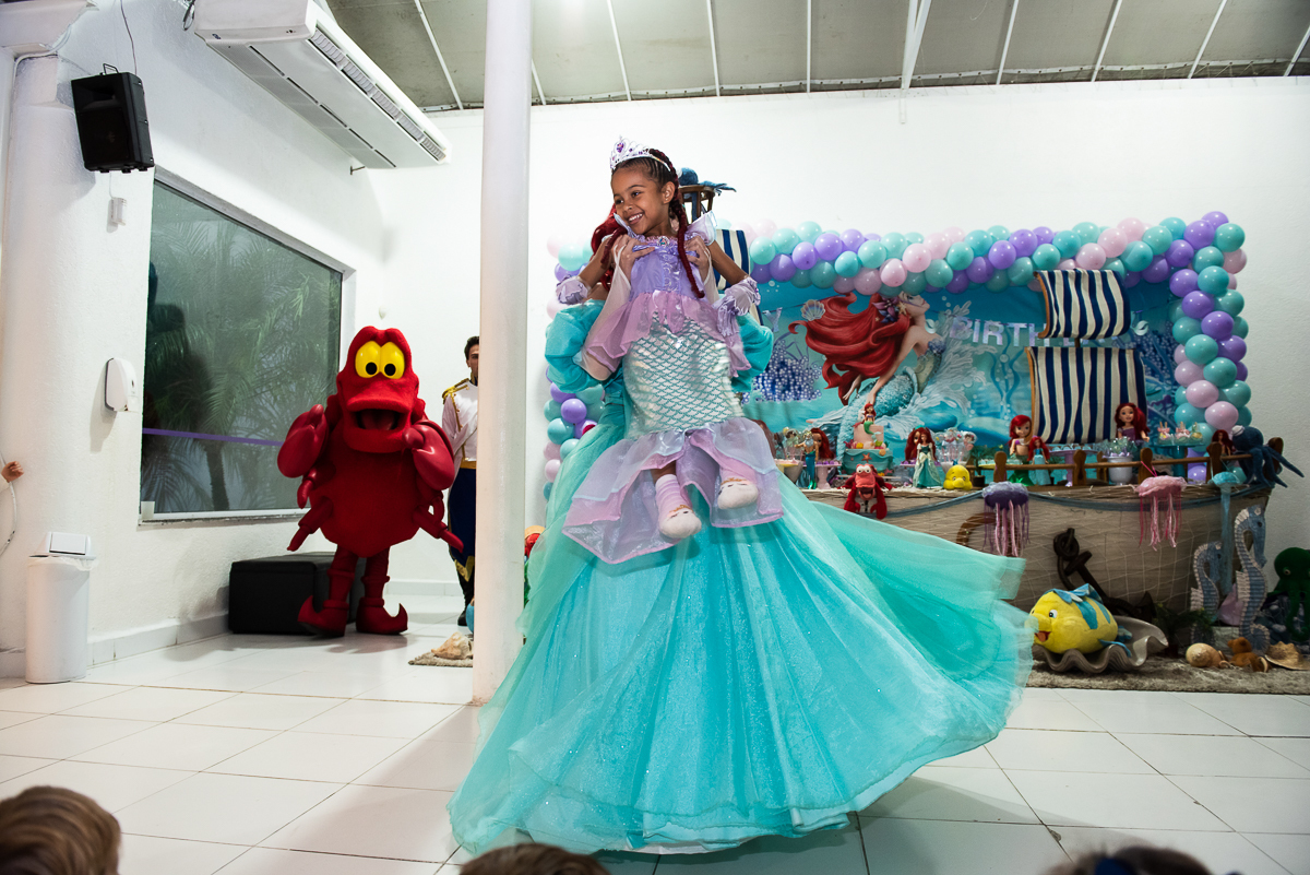 Moça de vestido azul pegou no colo a menina de vestido lilás