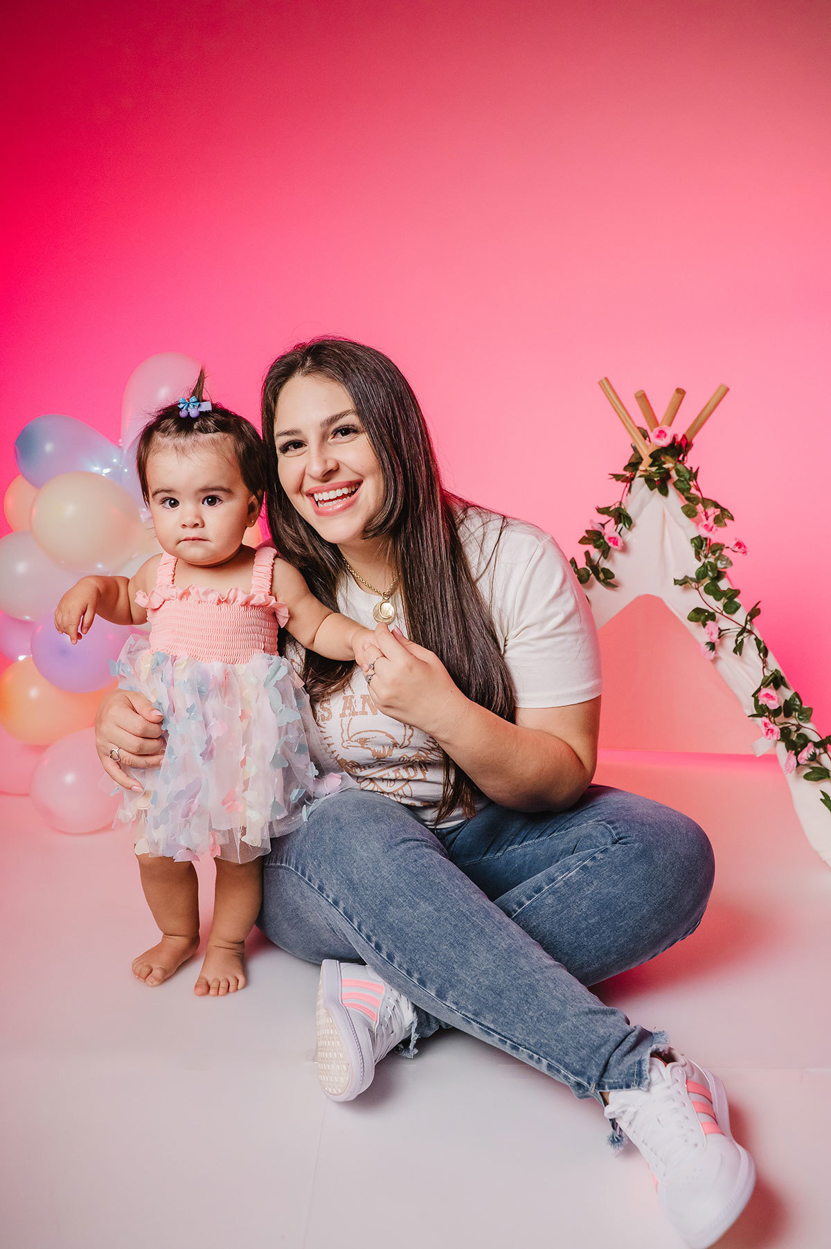 Foto da Lara com a mamãe no estúdio fotográfico, com bexigas e cabaninha com flores ao fundo