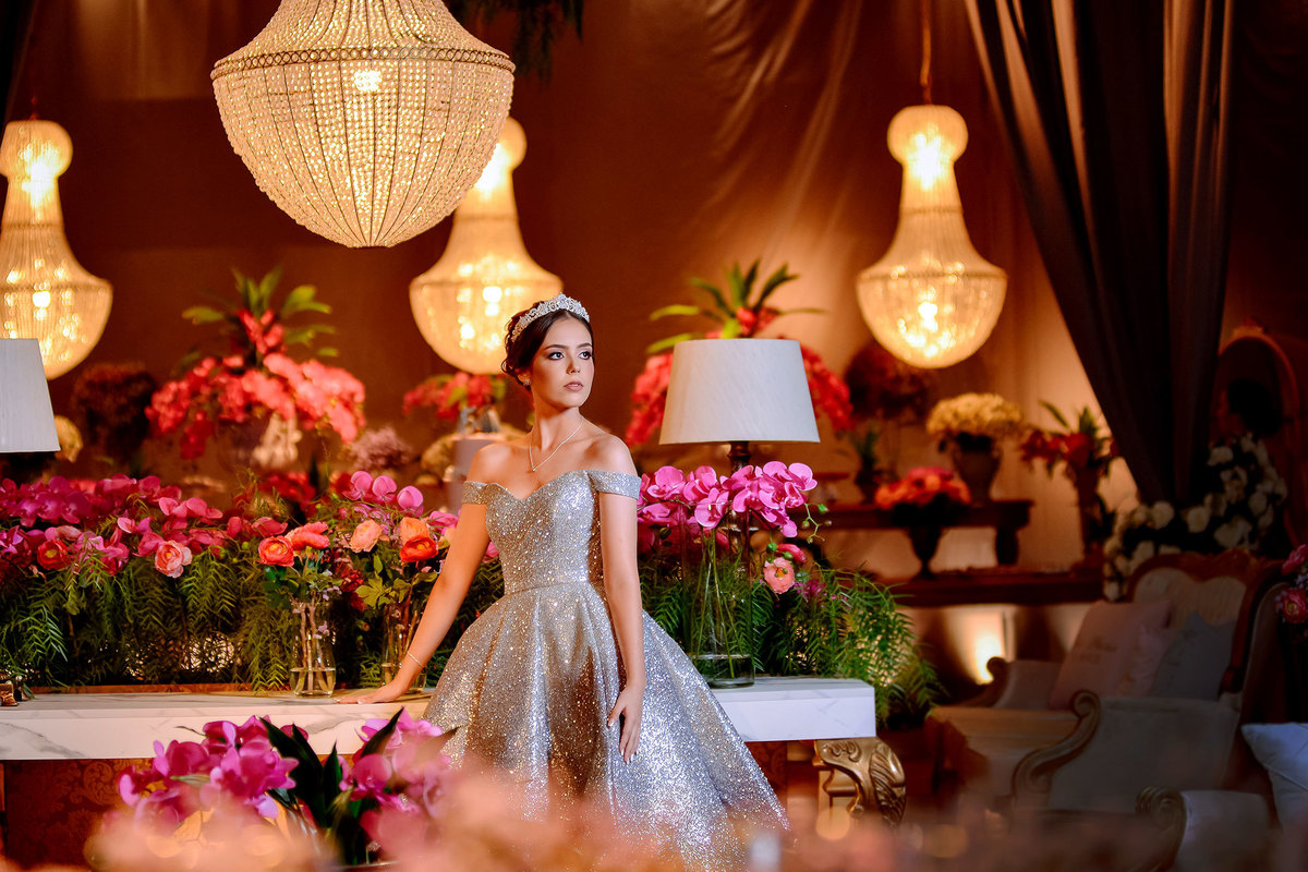 debutante em pé no meio da decoração do salão de festas, entre lustres e flores