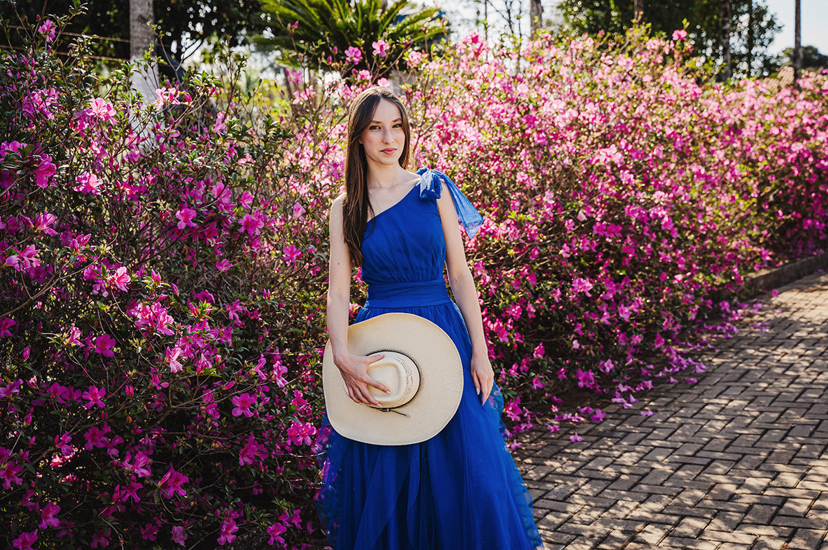 A aniversariante com vestido azul segurnando o chapeu com flores rosa ao fundo