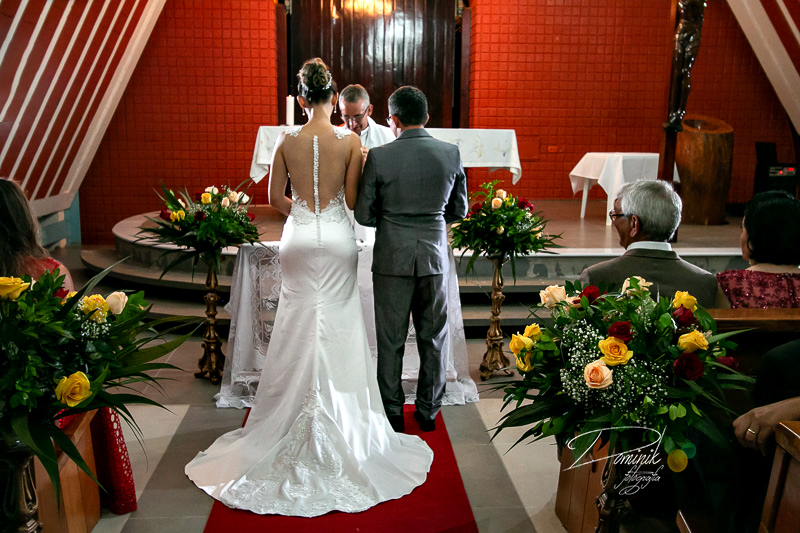 casal de noivos dentro da igreja de costas para o fotografo, flores na decoração tapete vermelho vestido branco terno cinza padre ao fundo casamento celebração terra nova itauba matupá sinop sorriso  capela   fotografia criativa dominik lago de matupa
