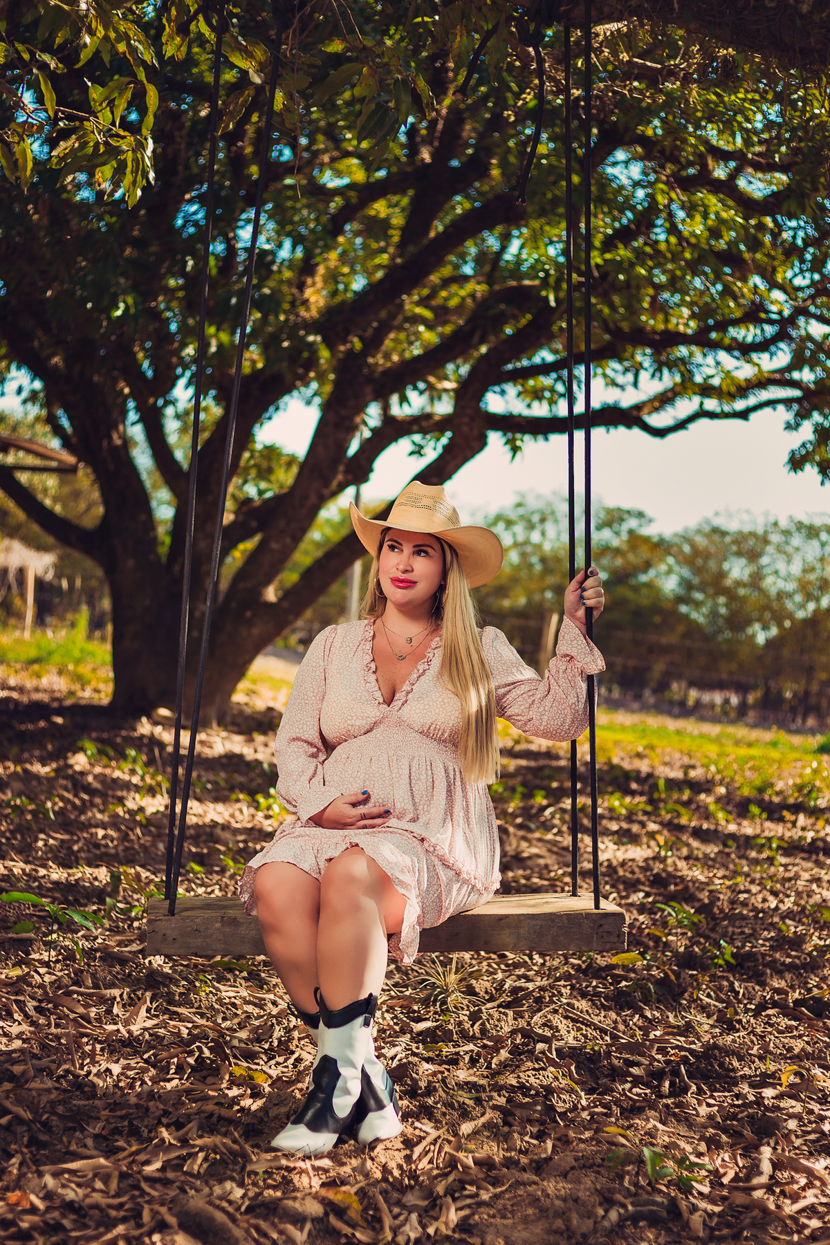 gestante sentada no balanço com um chapéu na cabeça em uma fazenda 
