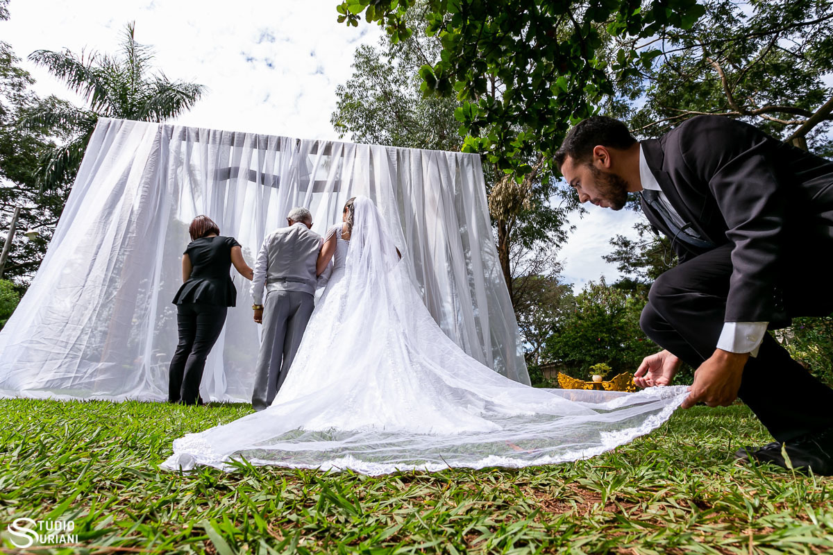 Cerimonial arrumando o vestido da noiva antes da sua entrada no casamento atrás de um véu para não ser vista pelo noivo no altar  