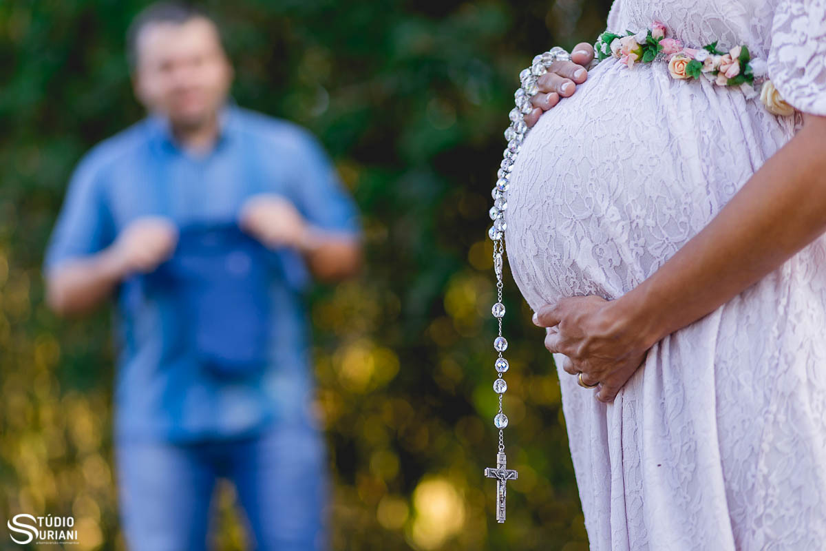 Pai segurando roupinha do bebê e mamãe segurando o sando terço na barriga com flores cor de rosa  