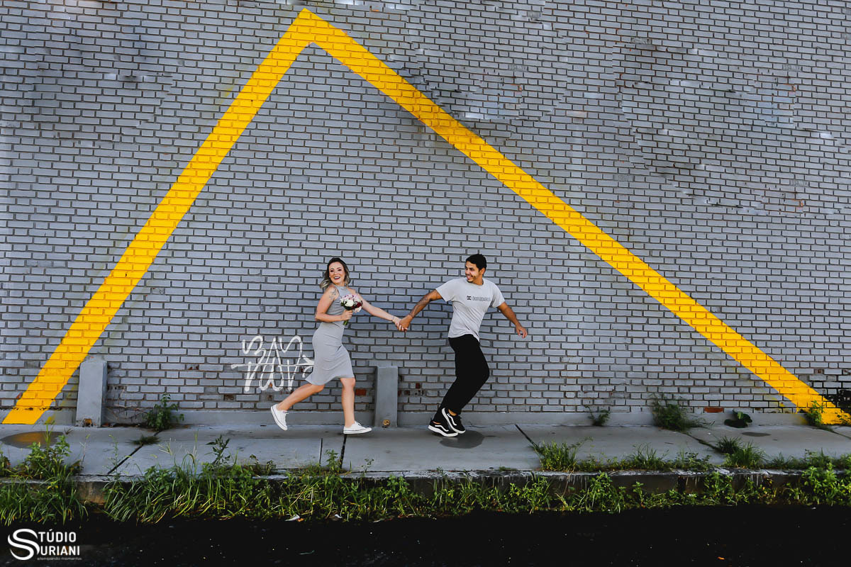 Fotografia criativa do fotógrafo Rogério Suriani, noivos correndo em parede cinza com buque de flores e pintura geométrica pintada de amarelo 