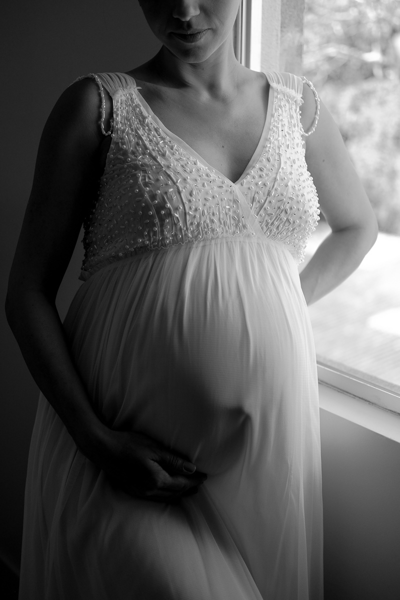 retrato em preto e branco feito pela fotografa isis vieira fotografia da gestante flavia estacio vestida com um lindo vestido branco do lado da janela de seu quarto no bairro corrego grande em florianopolis sc