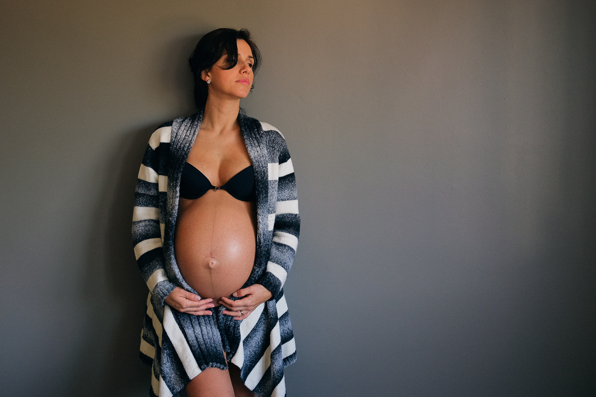 a gestante flavia posa encostada em uma parede toda pintada de cinza na sala da sua casa num registro do seu ensaio de gestante realizado em florianopolis sc