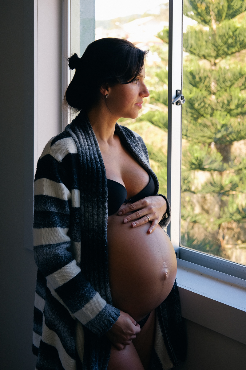 na janela de sua casa a gestante flavia faz carinho na barriga em momento de amor fotografado por isis vieira fotografa de familias e  estas infantis em florianopolis sc
