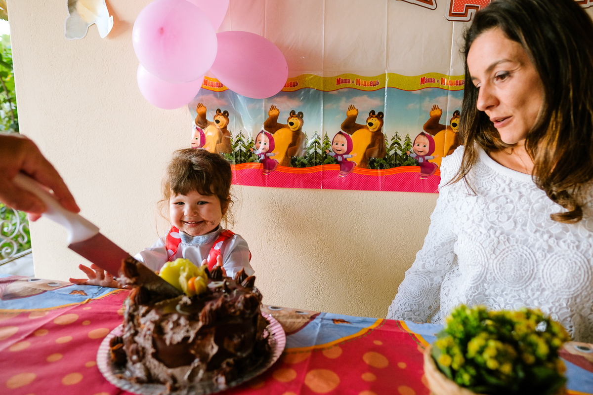 a criança olha com muita alegria o seu bolo de aniversário ser cortado para que ela possa come-lo na sua festa infantil realizada em casa em florianopolis na lagoa da conceição