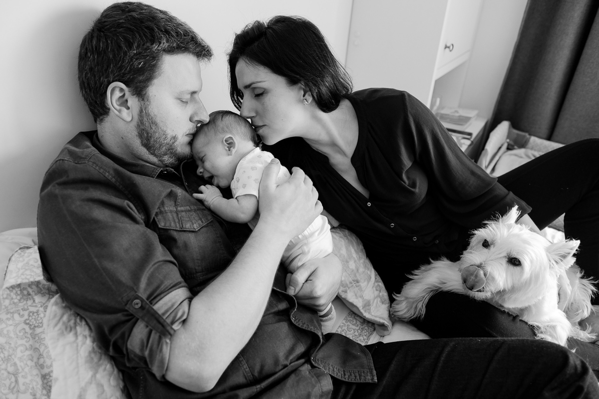 pai e mãe beijam o filho recém nascido na cabeça em registro de amor no ensaio fotografico de familia realizado em casa em florianopolis