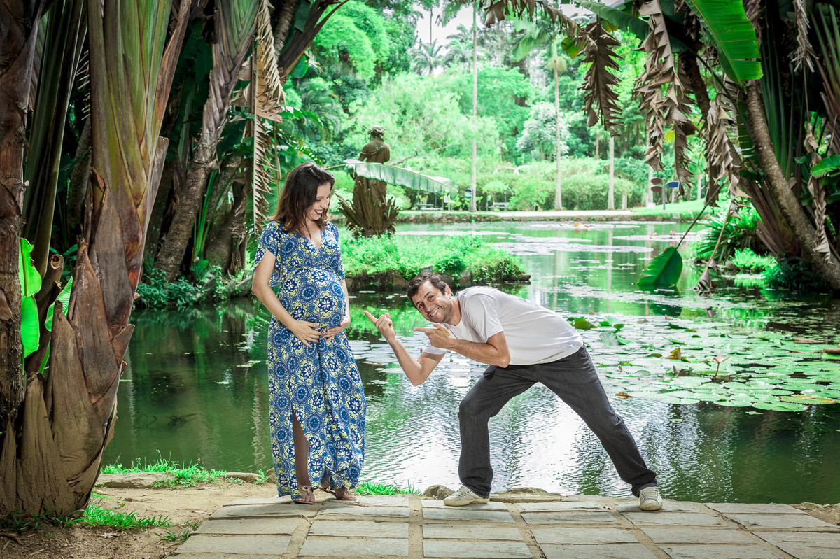 Papai apontado para barriga de gestante e sorrindo na beira de lago cercado por palmeiras em jardim botânico