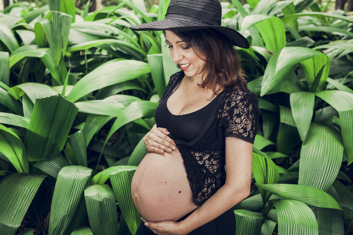gestante elegante e romantica segurando a barriga e usando chapeu em meio a floresta