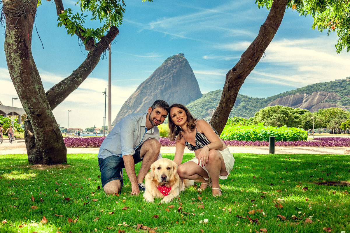 casal posando para foto com pet labrador embaixo de árvores no aterro do flamengo com pão de açúcar ao fundo