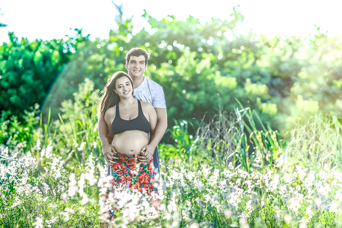 Pai e Gestante abraçados e sorrindo em lindo campo de flores com só em contra luz