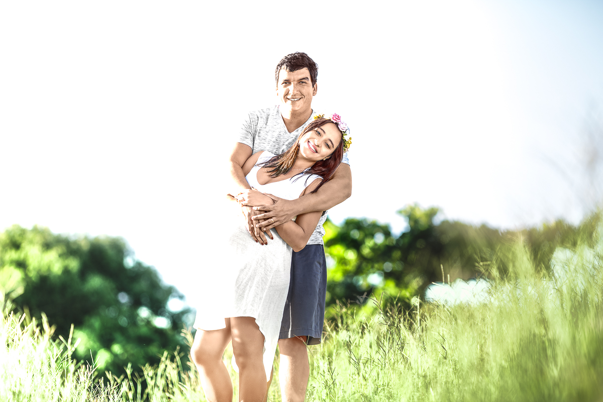 Pai e Gestante sorrindo e abraçados em lindo campo verde