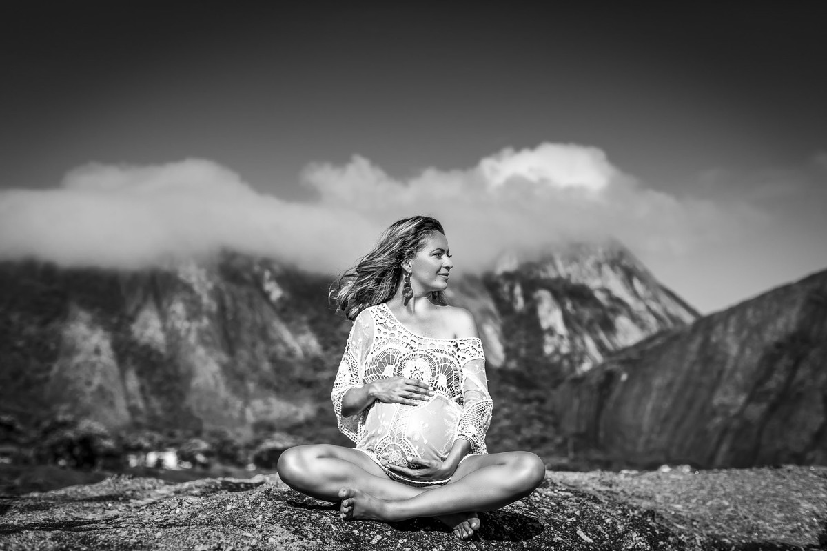 Gestante loira de vestido branco segurando a barriga com as pernas cruzadas na praia com montanhas ao fundo