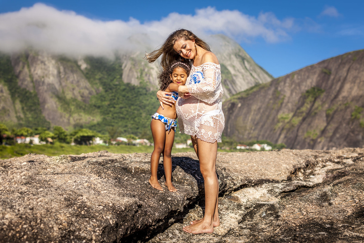 Gestante mulher loira recebendo carinho da filha em itaquatiara com céu azul e montanha ao fundo