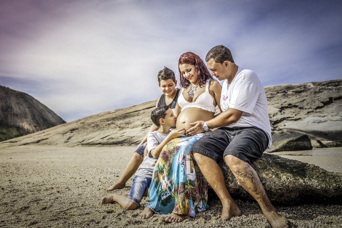 família e gestante sentada na pedra segurando a barriga e filho caçula beijando a barriga na praia de itaquatiara