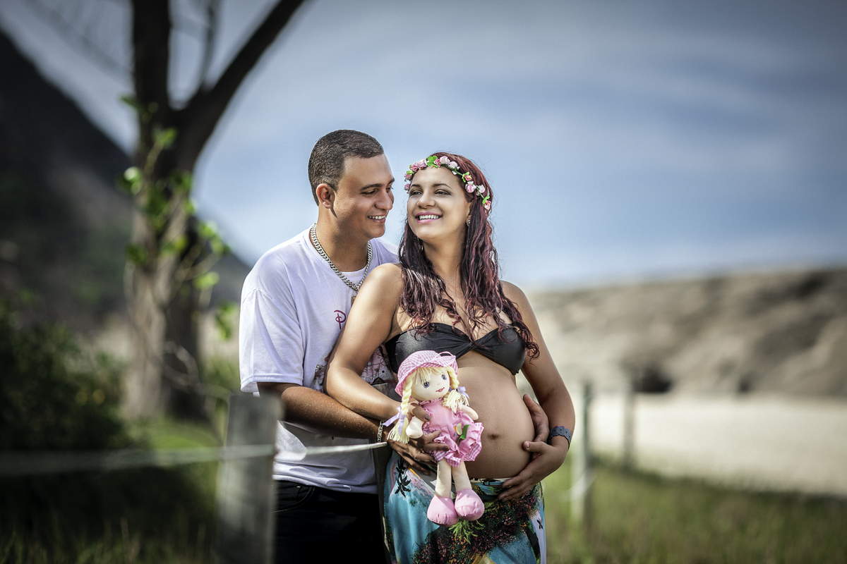Gestante e Papai sorrindo segurando uma bonequinha de panoe a barriga e sorrindo junto a uma cerca de arame e madeira na praia de itaquatiara