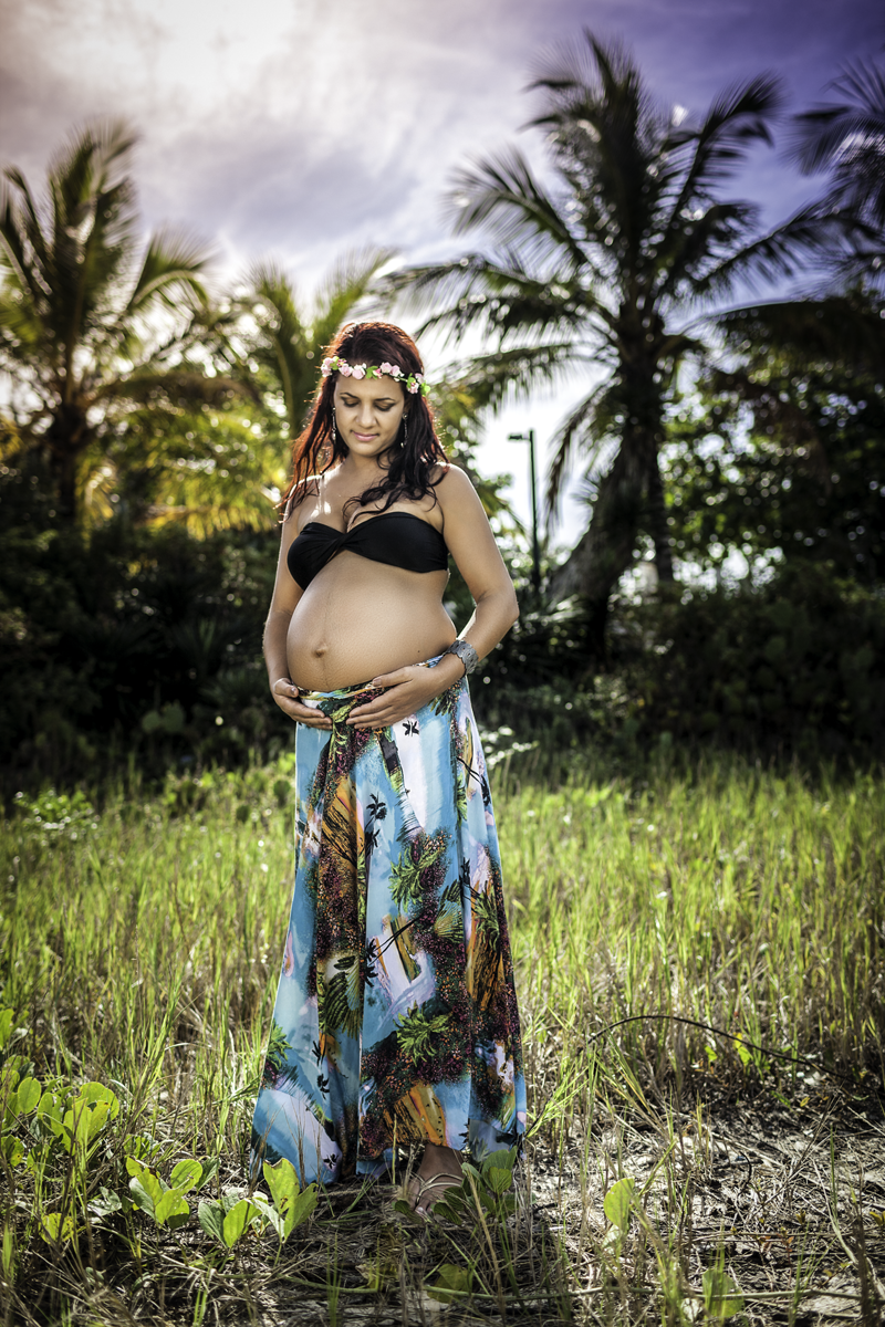 Gestante vestindo saia colorida top preto e coroa de flores segurando barriga em gramada verde com árvores e palmeira ao fundo em contra luz no nascer do sol segurando barriga e olhando para barriga