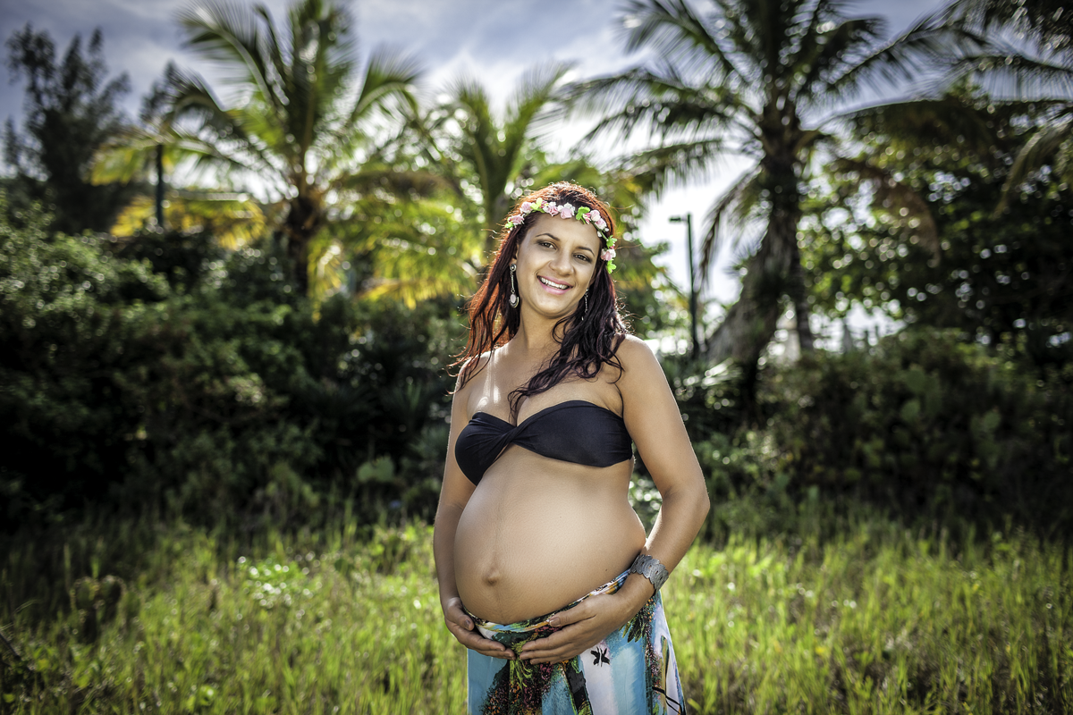 Gestante vestindo saia colorida top preto e coroa de flores segurando barriga em gramada verde com árvores e palmeira ao fundo em contra luz no nascer do sol segurando a barriga e sorrindo