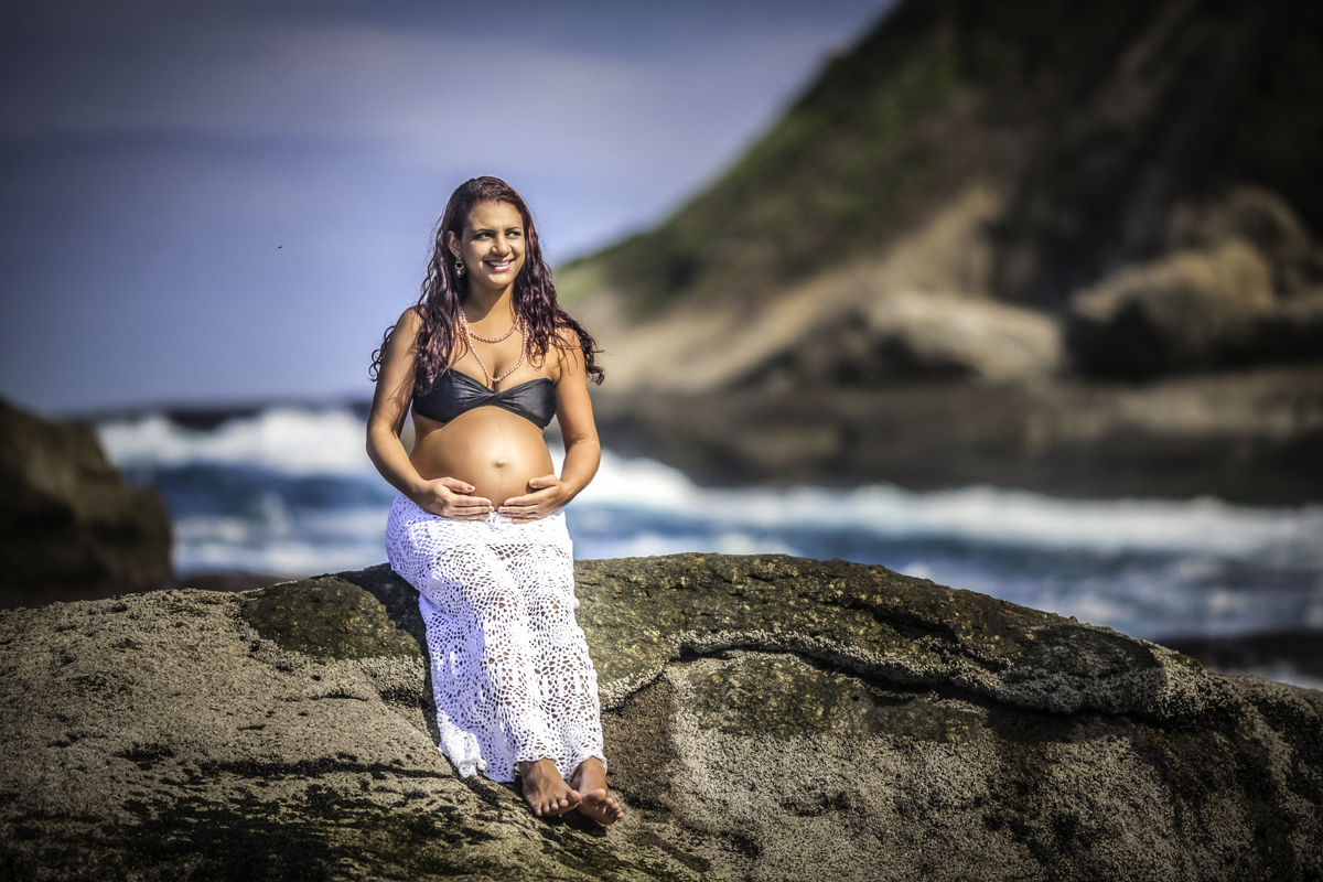 gestante vestindo saia branca de renda com top preto segurando a barriga em cima de pedra com o mar e montanha ao fundo
