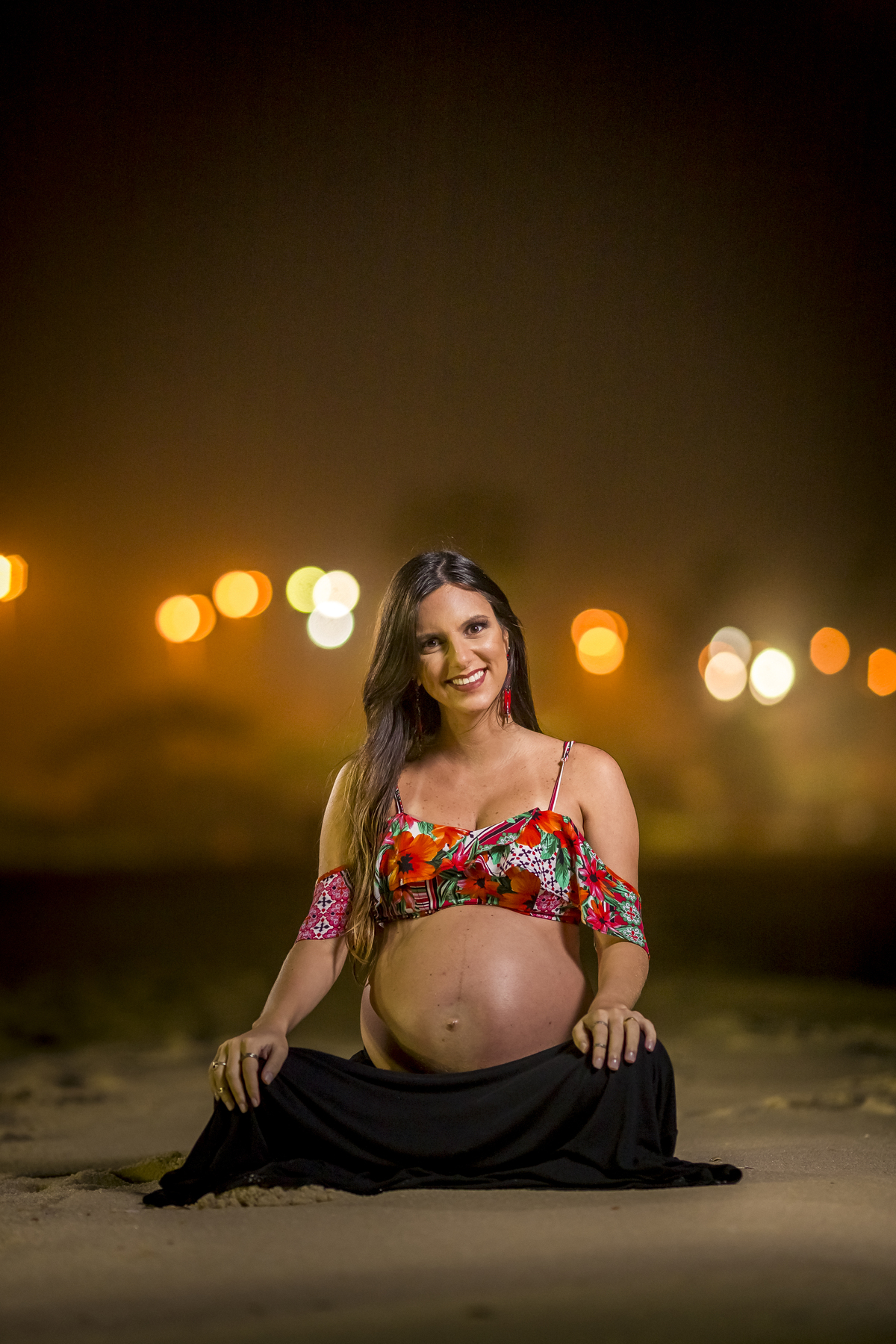 Gestante morena posando para ensaio noturno na praia do recreio sorrindo e segurando a barriga sentado